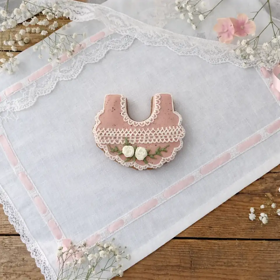Bolacha de especiarias decorada em glacê real, formato babador rosa com renda branca e flores, ideal para chá de bebê.